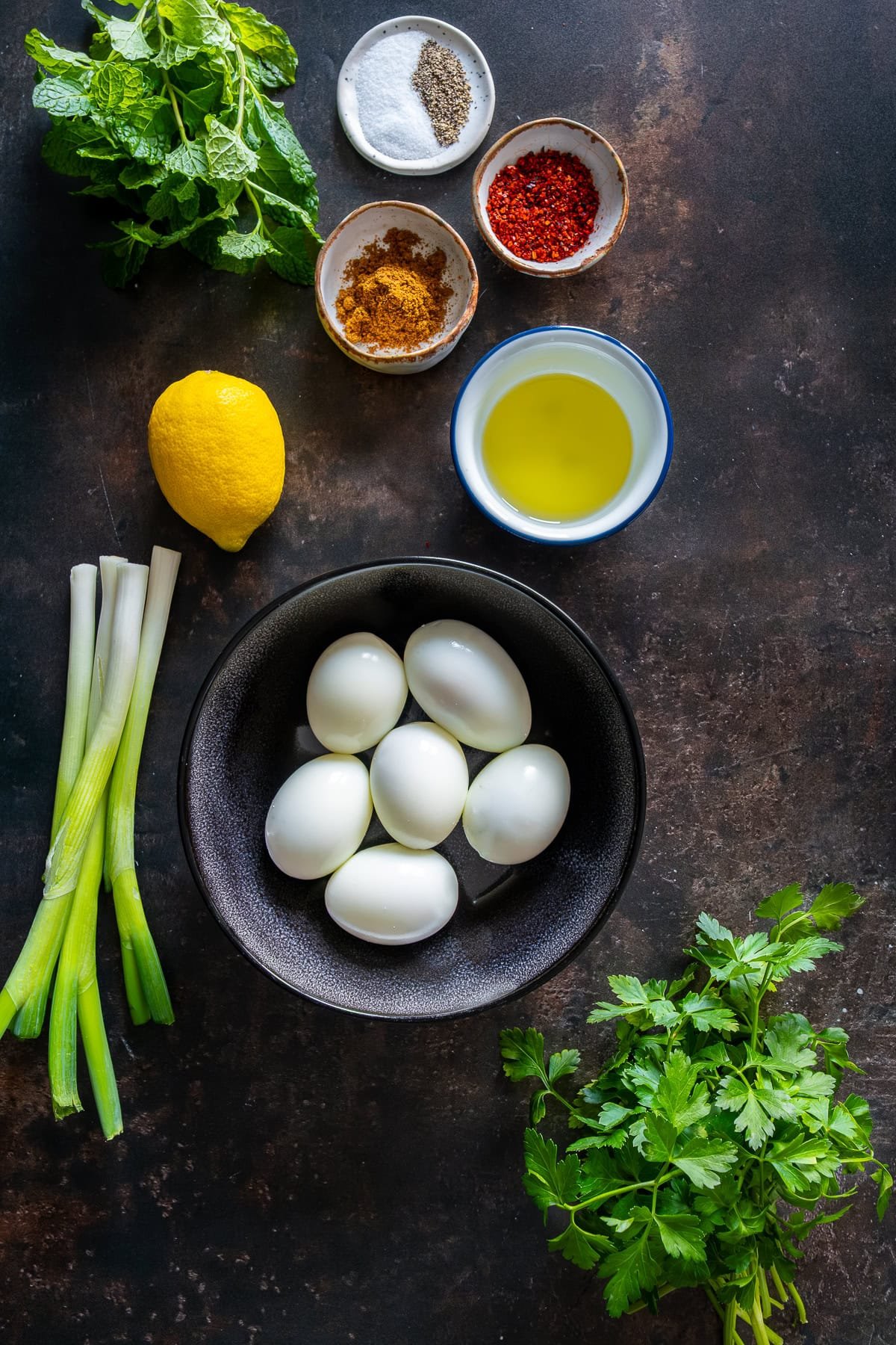 Overhead view of ingredients for Turkish egg salad including boiled eggs, lemon, olive oil, cumin, pul biber, fresh mint, parsley, and green onions.
