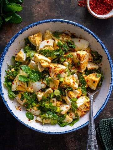 Overhead view of Turkish egg salad with boiled eggs, fresh herbs, and spices in a white bowl on a dark background.