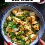 Overhead view of Turkish egg salad with fresh herbs and spices in a bowl, held by hands with a spoon.