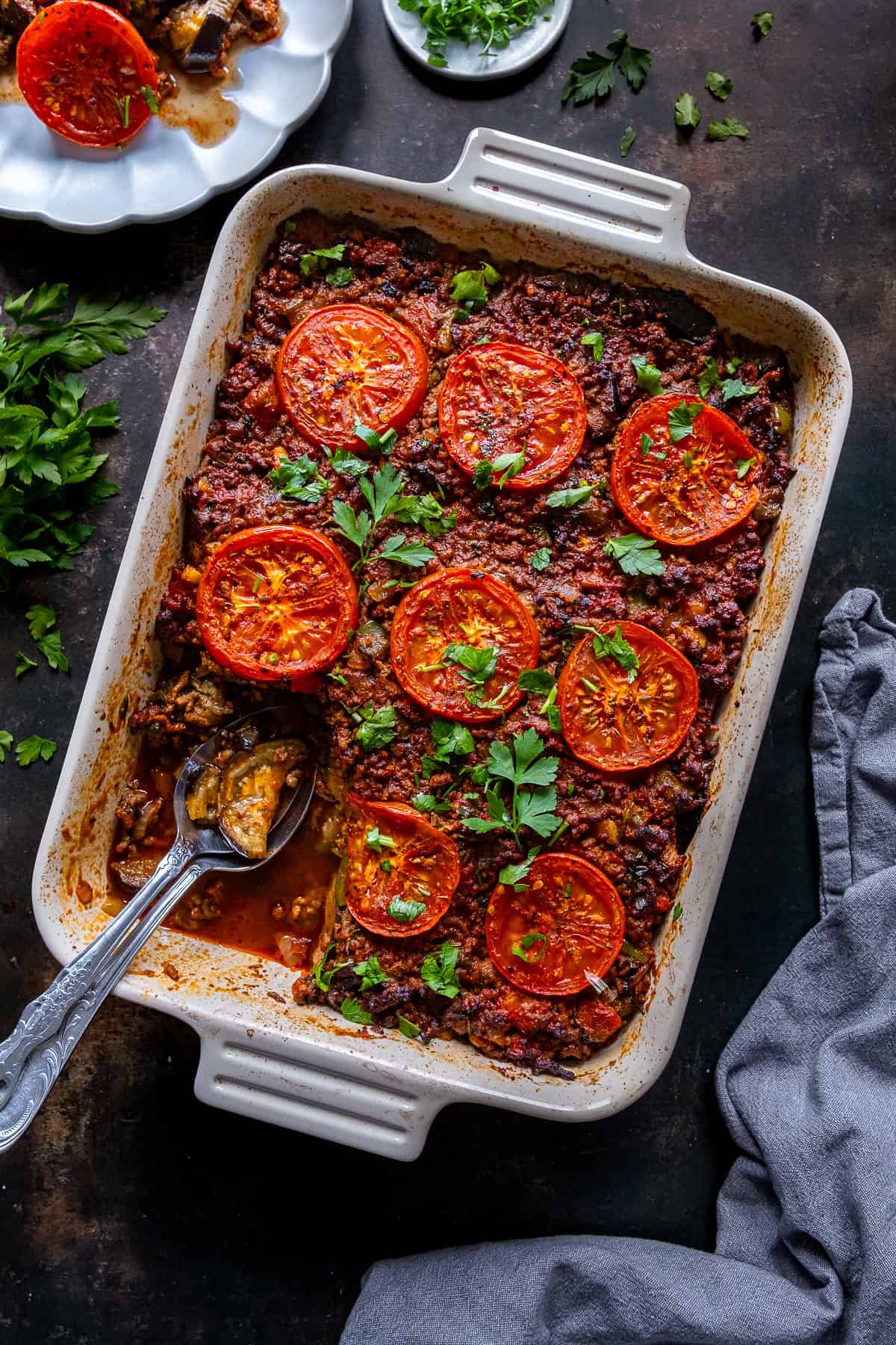Turkish moussaka in a baking dish topped with tomato slices and parsley, showing roasted eggplant and rich ground beef sauce.