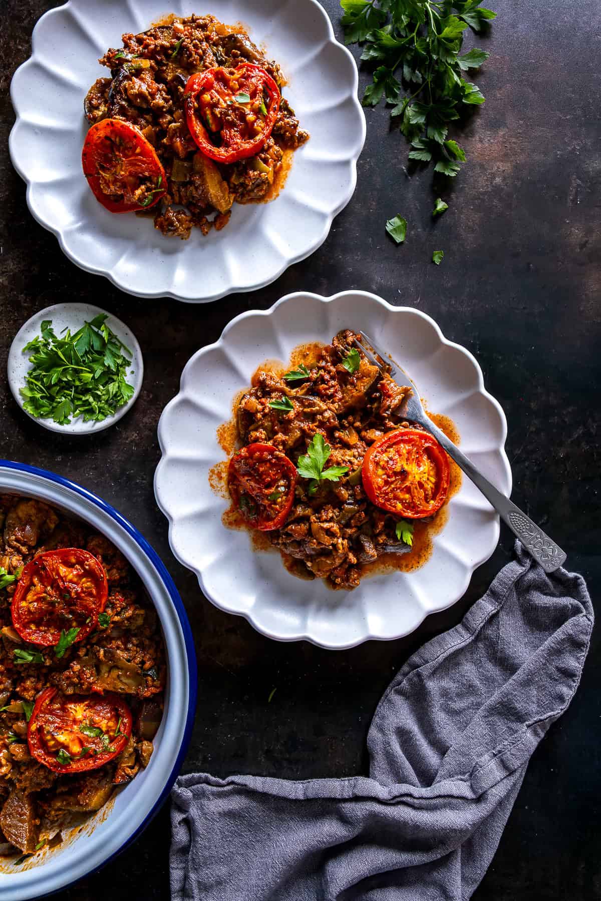 Two plates of Turkish moussaka topped with roasted tomato slices and parsley, served from a baking dish.