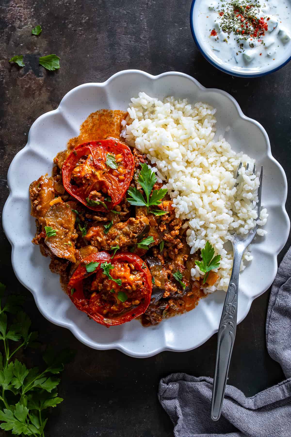 Plate of Turkish moussaka served with white rice and cacık, topped with roasted tomato slices and fresh parsley.