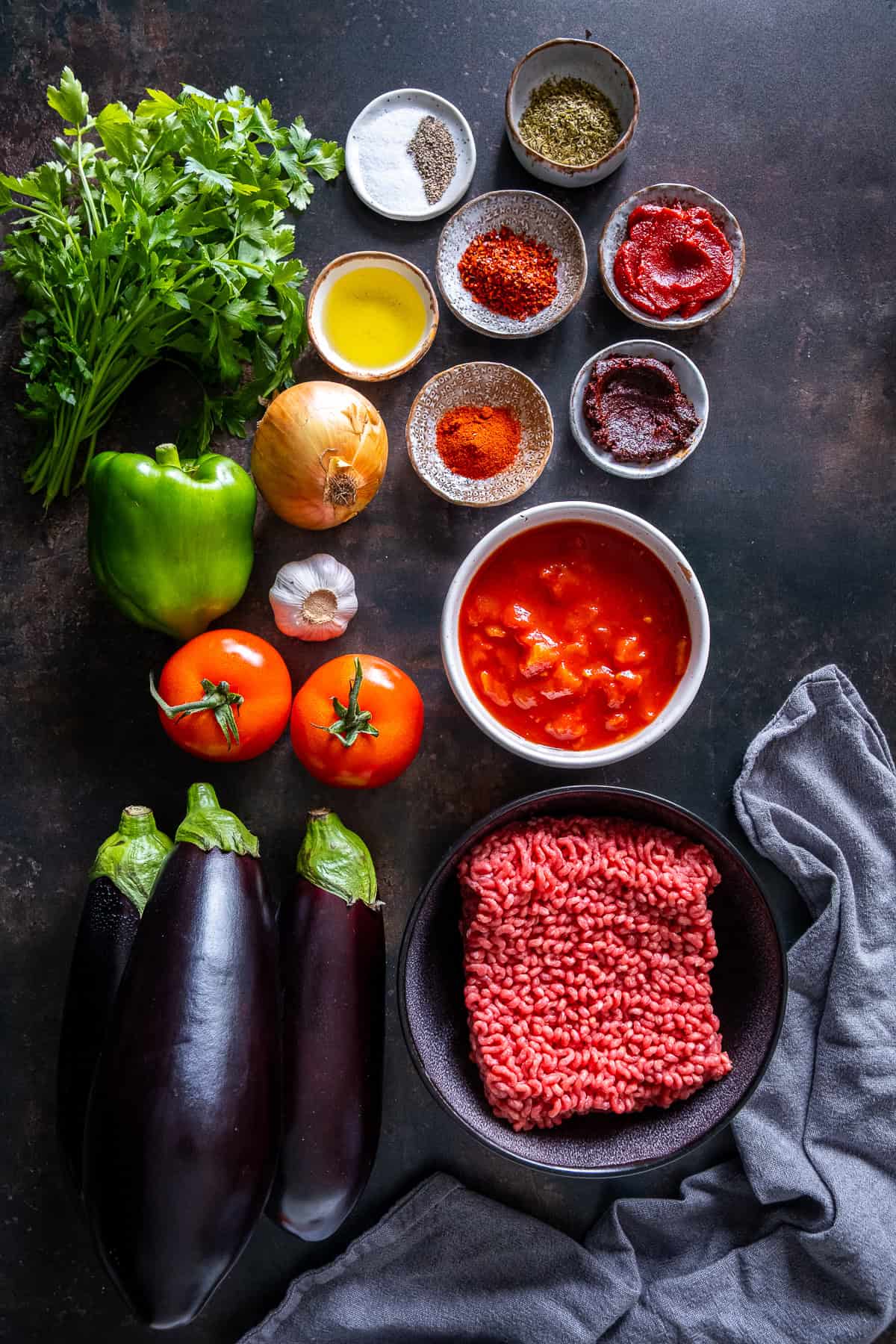 Ingredients for Turkish moussaka including eggplants, ground beef, tomatoes, onion, garlic, green pepper, olive oil, spices, and tomato paste.