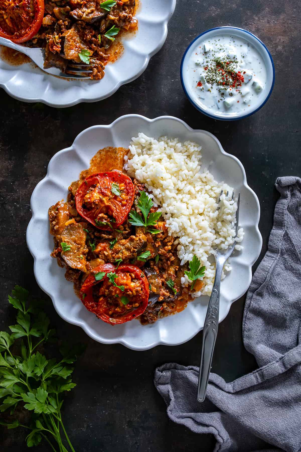 Plate of Turkish moussaka with white rice pilaf and cacık, topped with roasted tomato slices and fresh parsley.