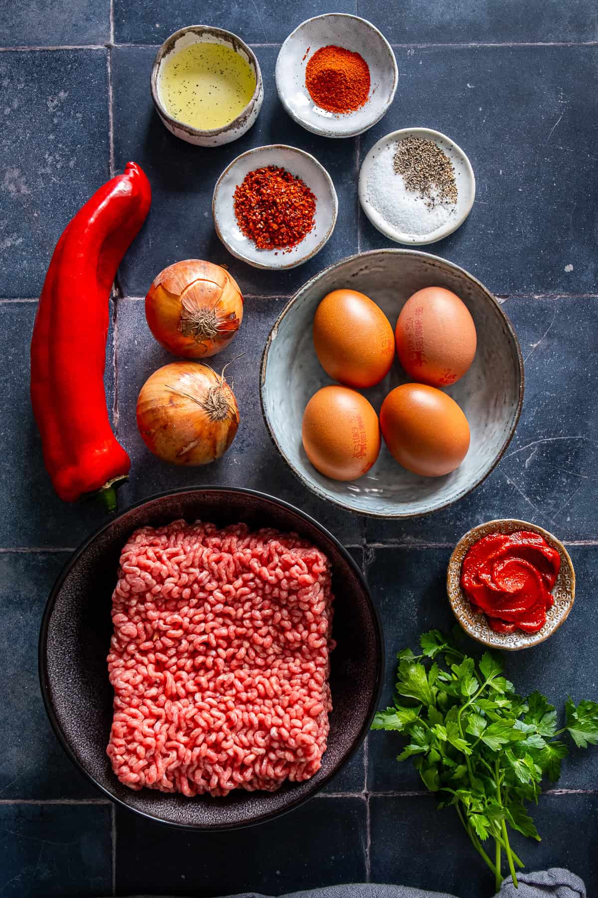 Ground beef, eggs, onions, red pepper, tomato paste, olive oil, parsley, and spices arranged on a dark surface as ingredients for ground beef and eggs breakfast.
