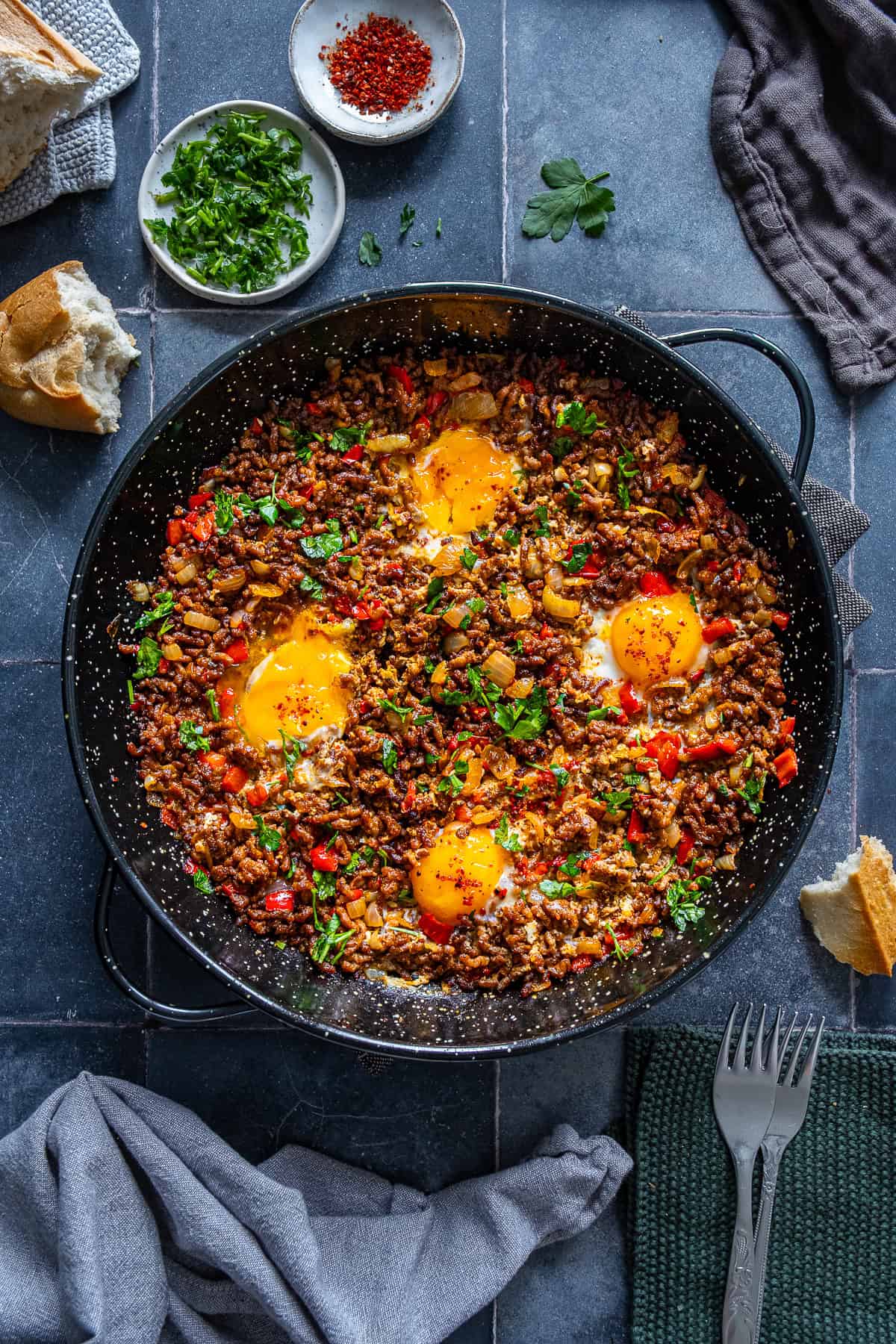 Ground beef and eggs cooked in a skillet with onions, red peppers, and parsley, topped with soft egg yolks and served with bread on the side.