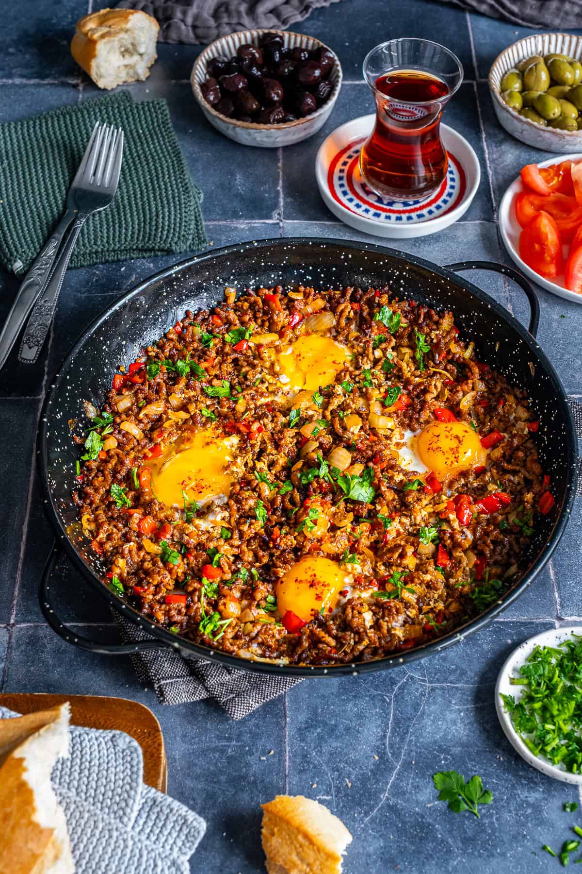 Ground beef and eggs cooked with onions and red peppers in a skillet, served with olives, tomatoes, bread, and Turkish tea for a savory breakfast.