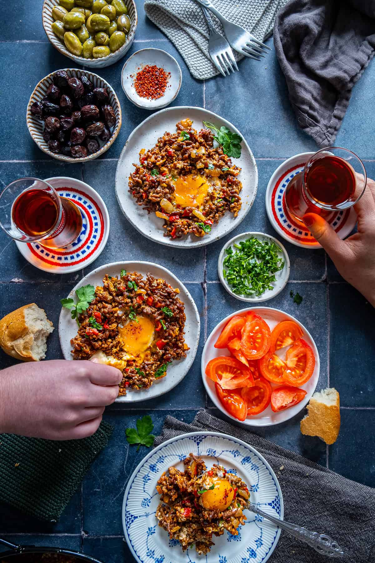 Hands dipping bread into egg yolk from ground beef and eggs while another person holds Turkish tea at a Turkish-style breakfast table with tomatoes and olives.
