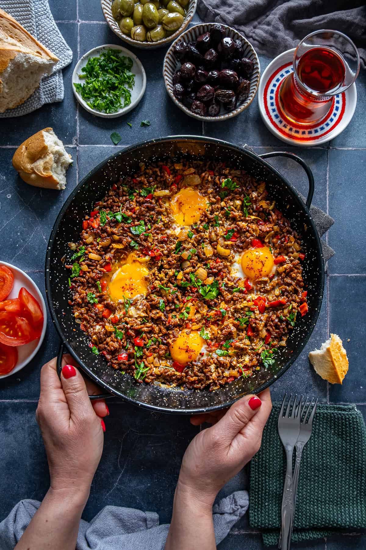 Hands holding a skillet filled with ground beef and eggs topped with parsley, surrounded by bread, olives, sliced tomatoes, and Turkish tea.