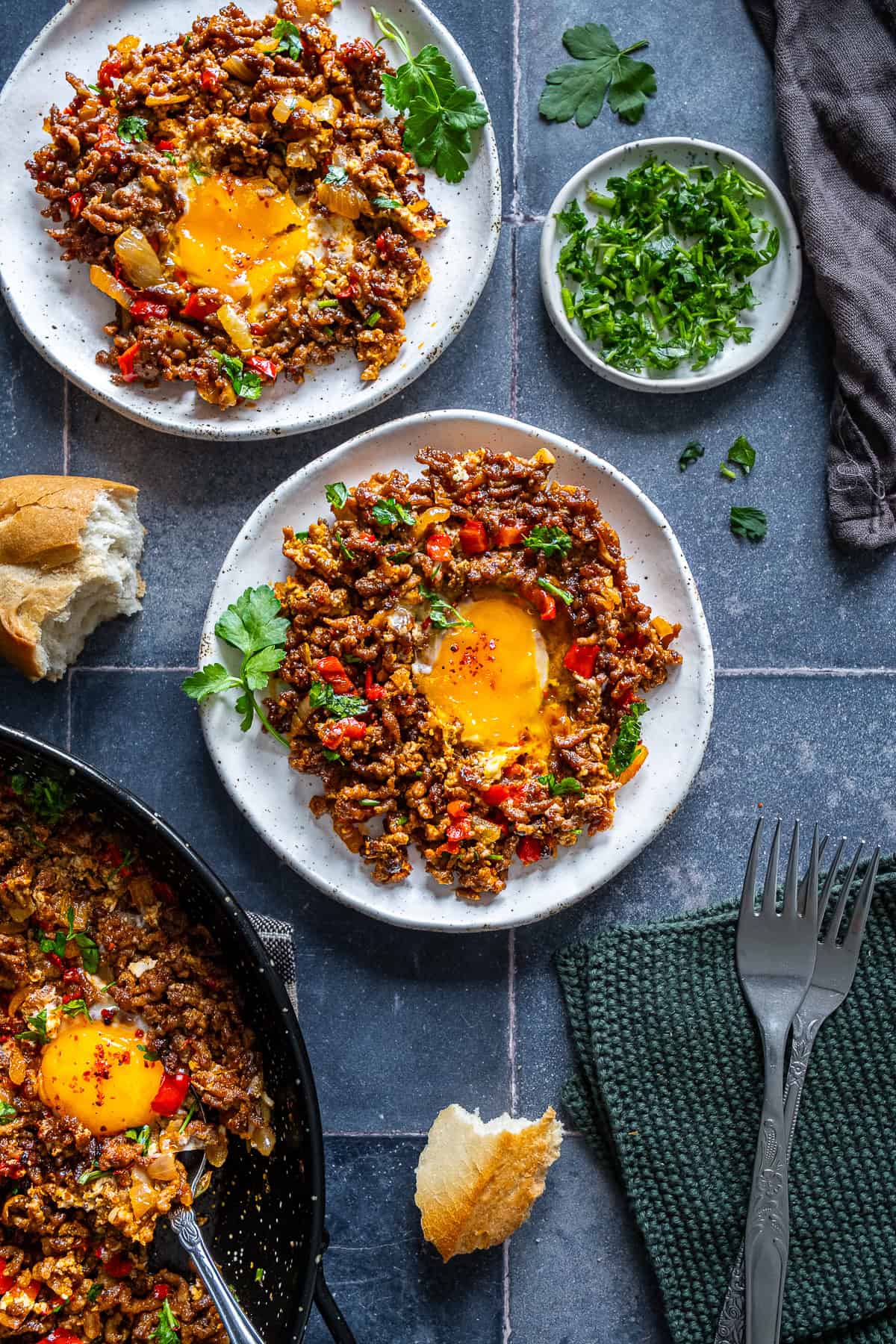 Plates of ground beef with soft egg yolks, cooked with onions and red peppers, served with bread and fresh parsley on the side.
