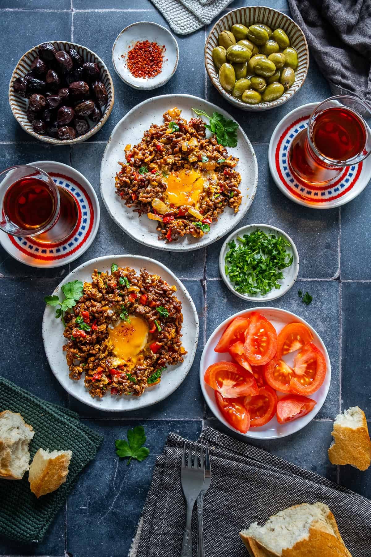Two plates of ground beef and eggs with runny yolks served with olives, sliced tomatoes, bread, and Turkish tea.