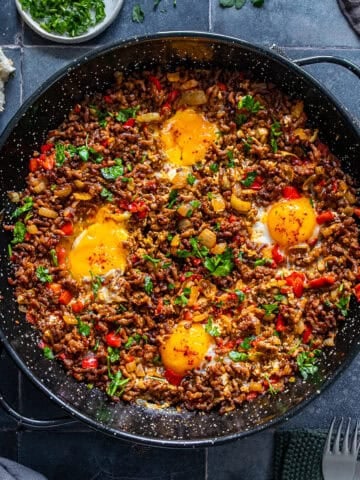 Ground beef and eggs cooked in a skillet with onions, red peppers, and parsley, topped with soft egg yolks and served with bread on the side.