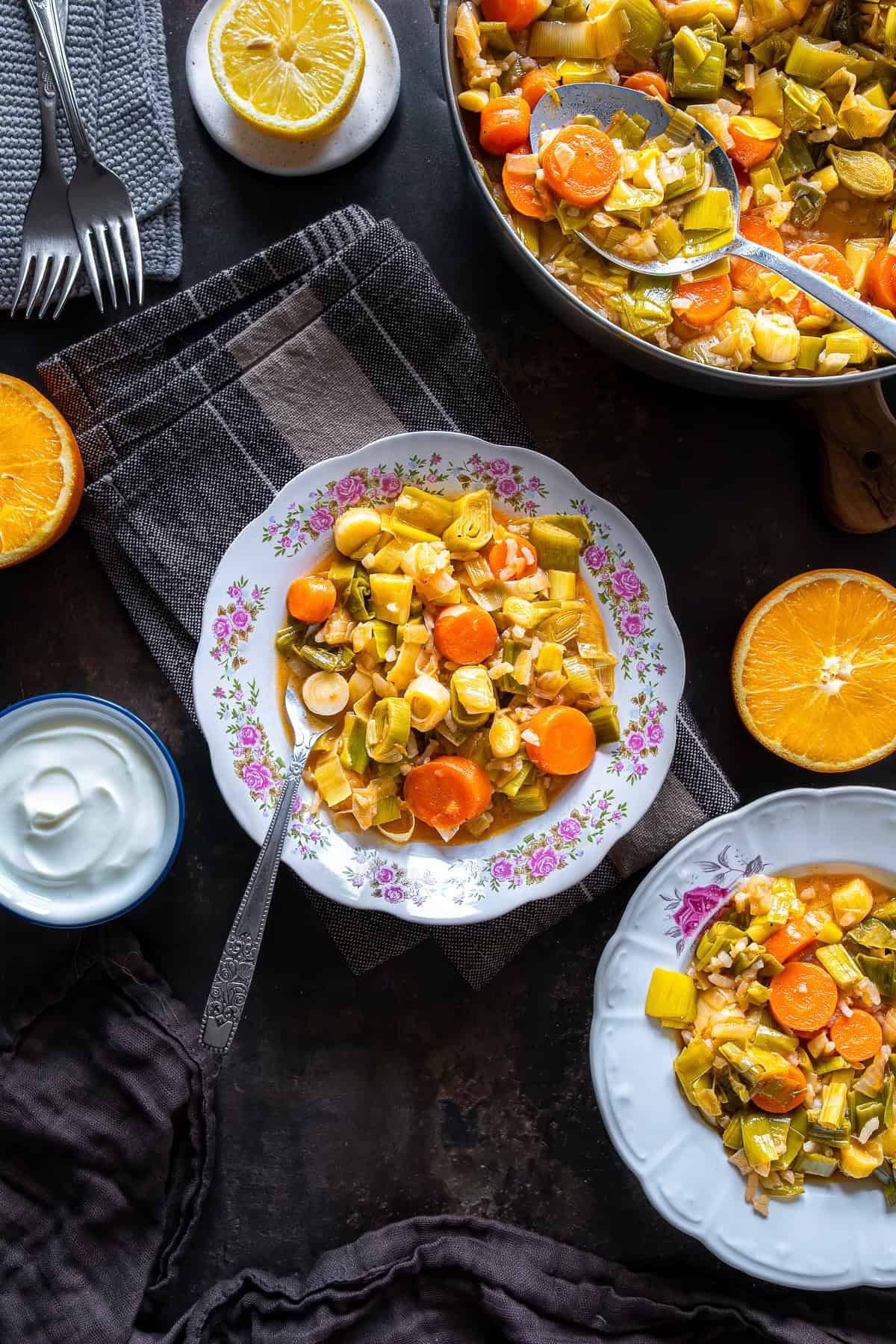 Overhead view of Turkish Leeks in Olive Oil with carrots and rice served on floral plates, alongside yogurt, lemon slices, and a pan of the dish on the table.