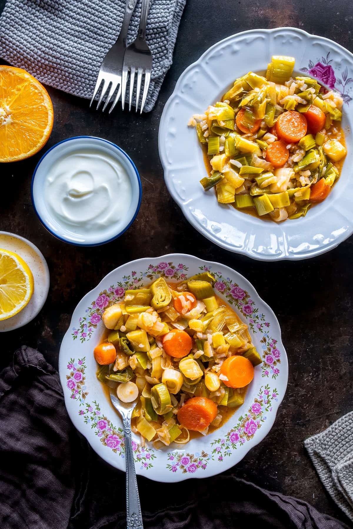 Top view of two plates of Turkish Leeks in Olive Oil with carrots and rice, served with a bowl of yogurt and lemon slices on a dark tabletop.