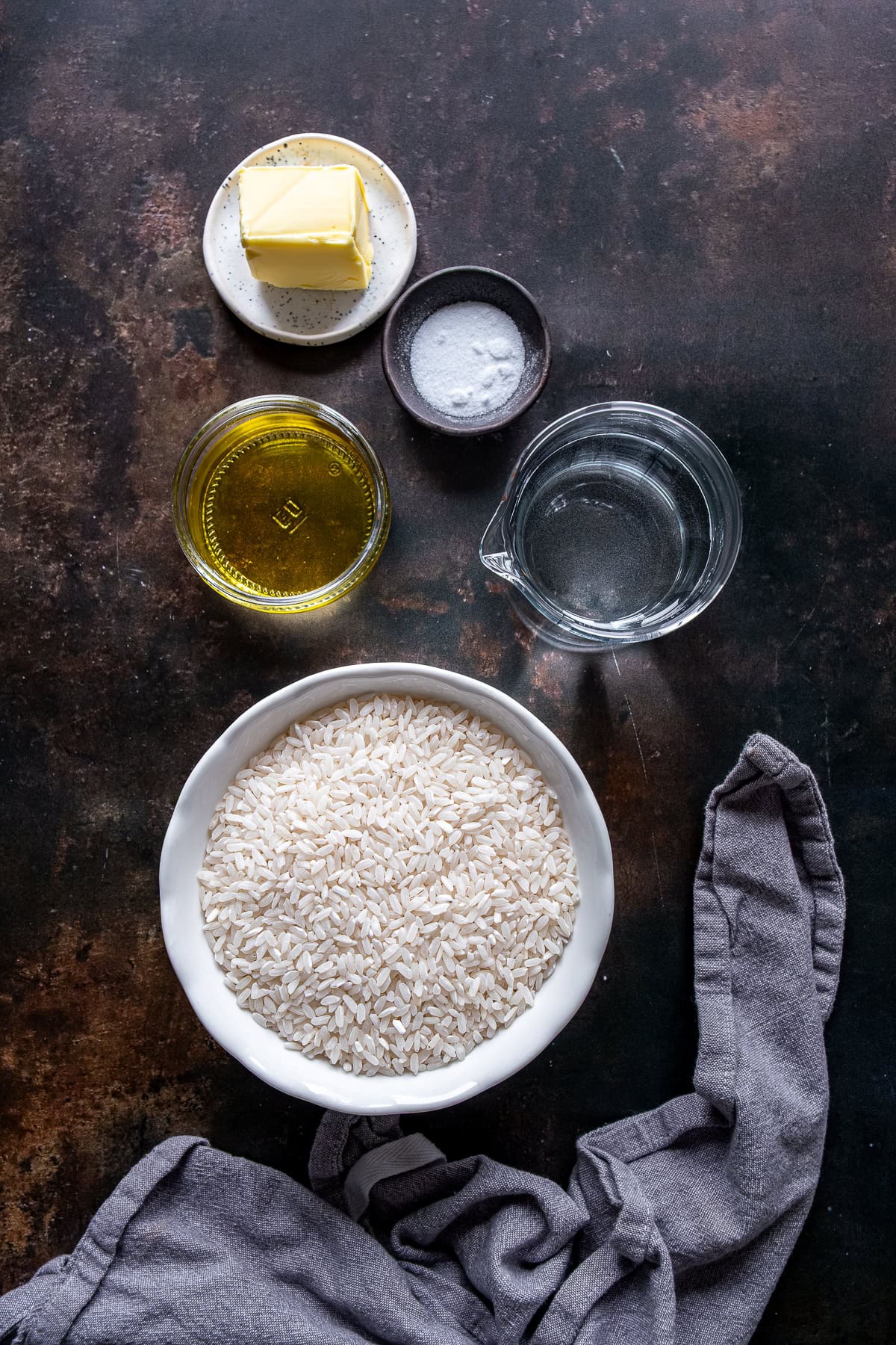 Overhead view of the ingredients for Turkish rice pilaf: uncooked rice in a bowl, olive oil, butter, salt, and water on a dark surface.
