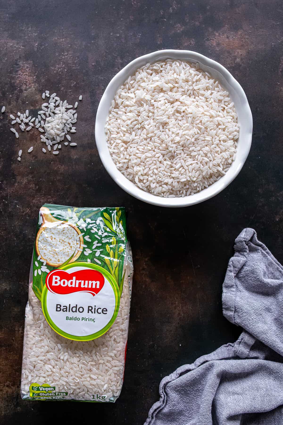 Bowl of uncooked Baldo rice with a package of Bodrum Baldo rice on a dark surface.