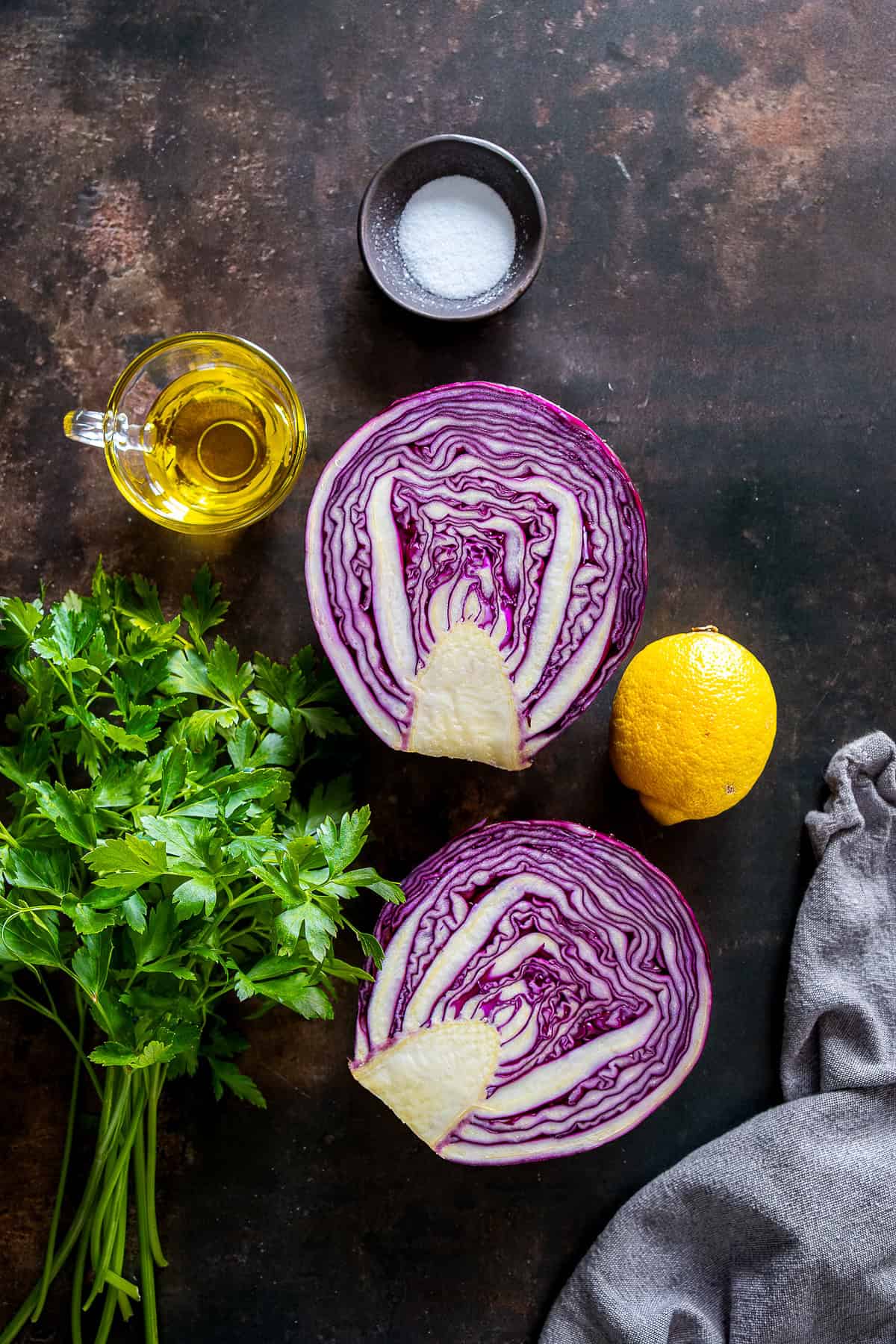 Halved red cabbage with parsley, lemon, olive oil, and salt on a dark surface.