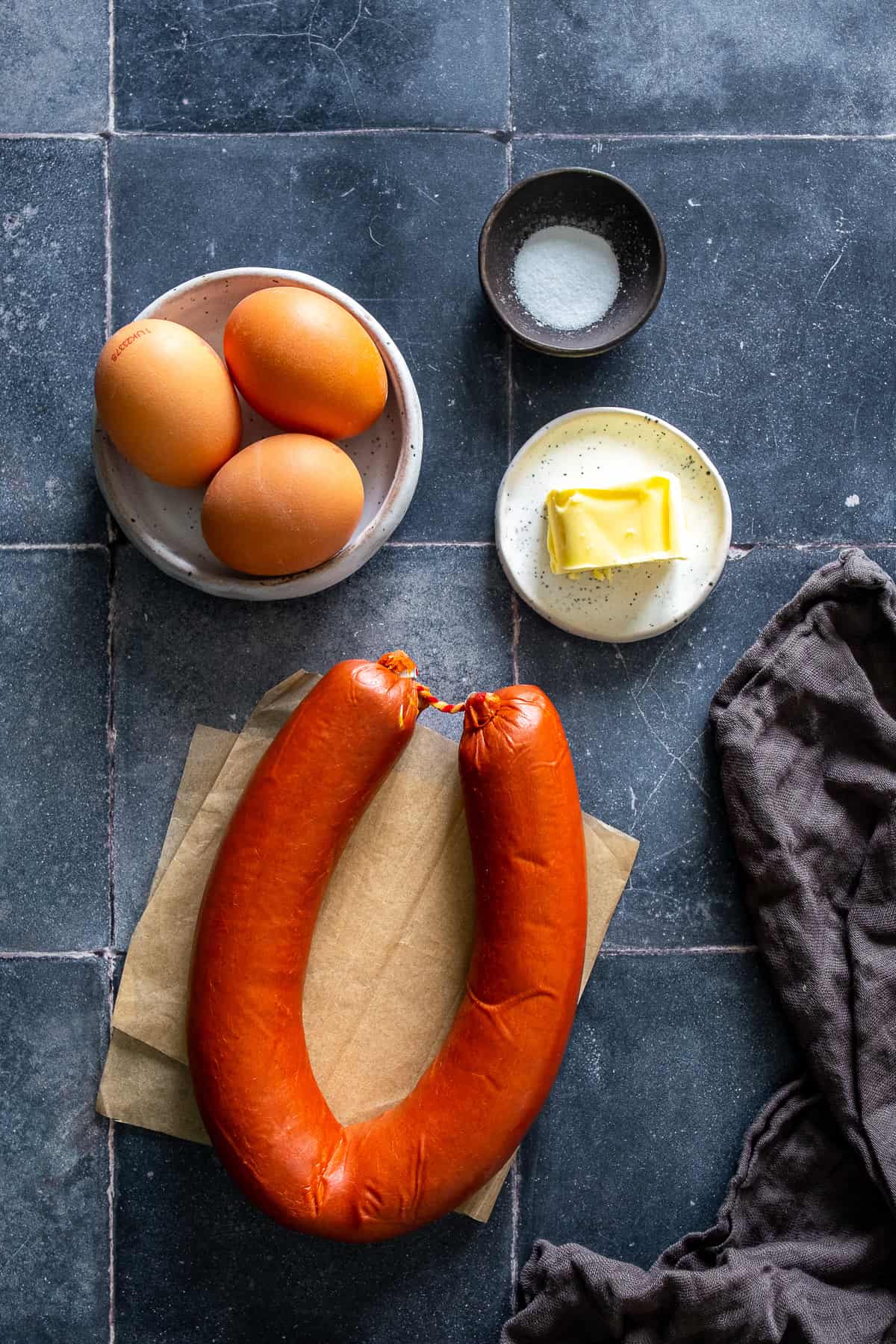 Ingredients for Turkish sucuk and eggs including Turkish sucuk, eggs, butter, and salt on a dark background.