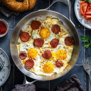 Overhead view of Turkish sucuk and eggs cooked in a pan and served with simit and tomatoes for breakfast.