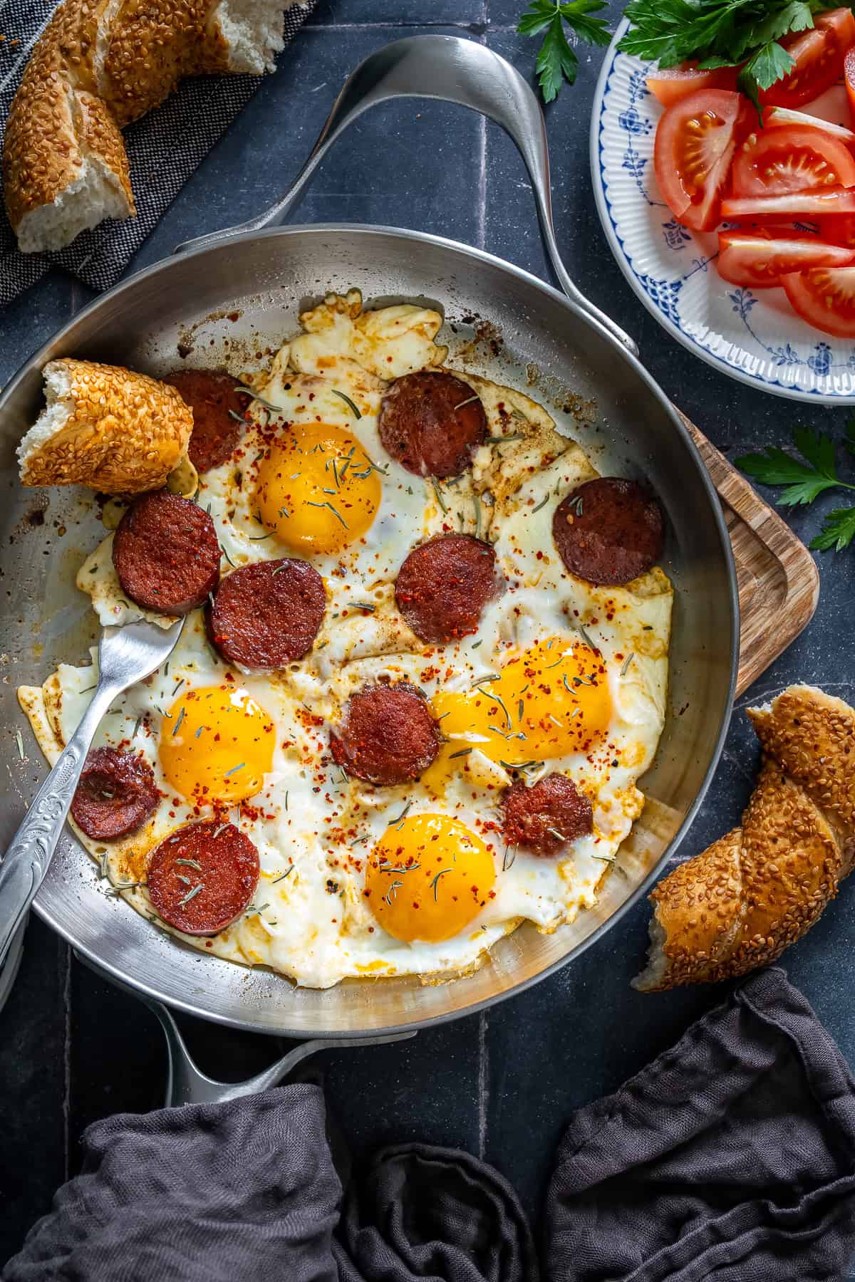 Turkish sucuk and eggs served in a pan with simit and sliced tomatoes on the side.