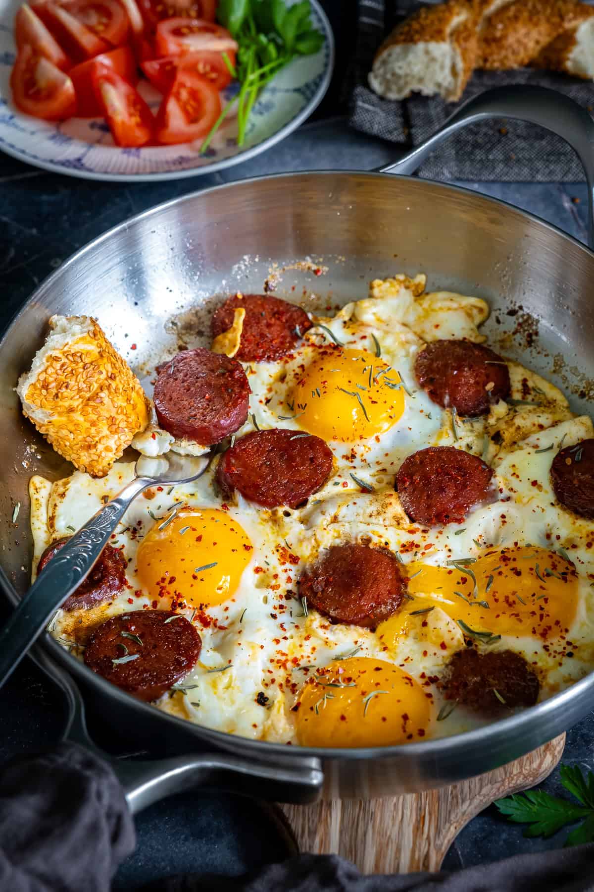 Close-up of Turkish sucuk and eggs in a pan with crispy sucuk slices, runny egg yolks, and simit on the side.