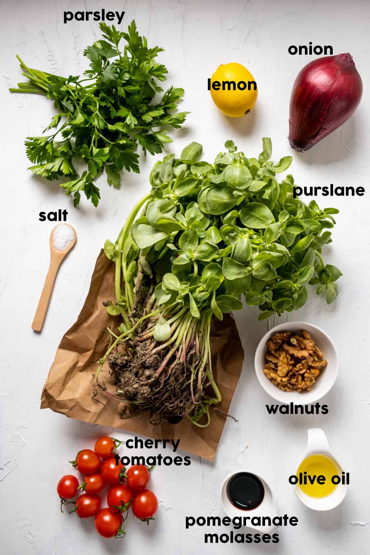 Ingredients for Turkish purslane salad arranged on a white surface, including fresh purslane with roots attached, parsley, red onion, lemon, cherry tomatoes, walnuts, olive oil, pomegranate molasses and salt.