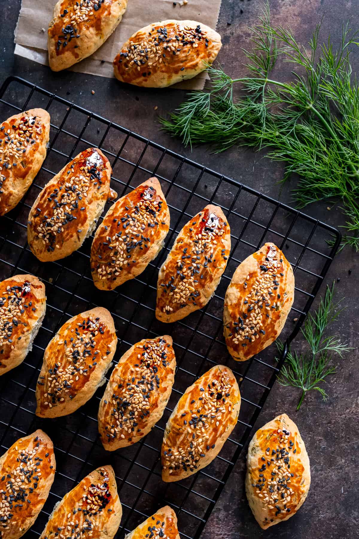 Freshly baked Turkish pogaca with dill (dereotlu pogaca) topped with sesame and nigella seeds, cooling on a wire rack with fresh dill sprigs.