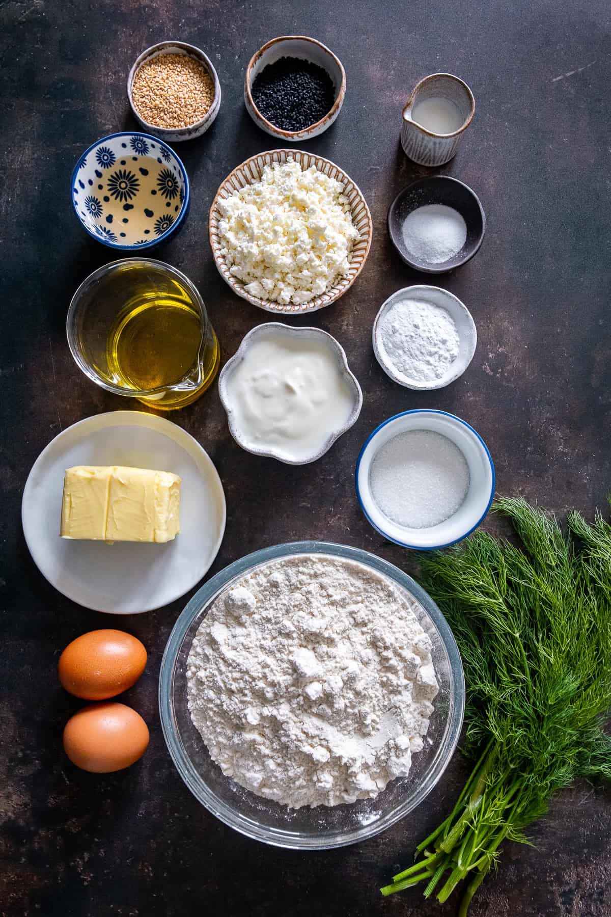 Ingredients for Turkish pogaca with dill (dereotlu pogaca), including flour, fresh dill, butter, yogurt, olive oil, eggs, feta cheese, baking powder, vinegar, sugar, sesame seeds, and nigella seeds.