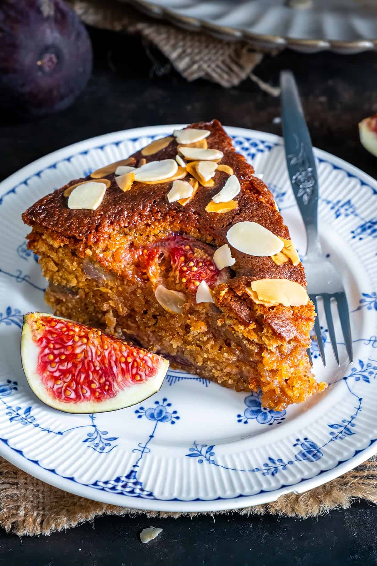 Close-up of a moist fresh fig cake slice topped with flaked almonds and served with a fresh fig.