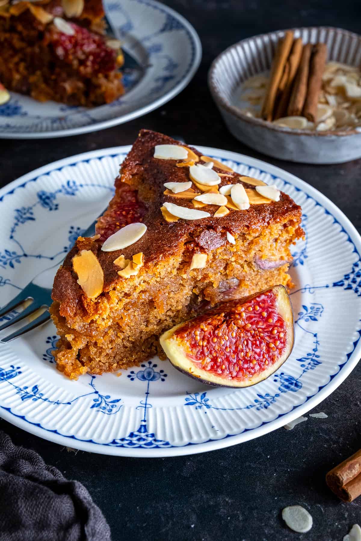 Thick slice of fresh fig cake with a soft, moist crumb and pieces of fig inside, topped with flaked almonds and served with a fresh fig half, with another slice and cinnamon sticks in the background.