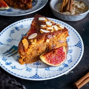 A slice of fresh fig cake with a thick, moist crumb and baked figs inside, topped with flaked almonds and served with a fresh fig half.
