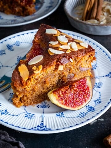 A slice of fresh fig cake with a thick, moist crumb and baked figs inside, topped with flaked almonds and served with a fresh fig half.