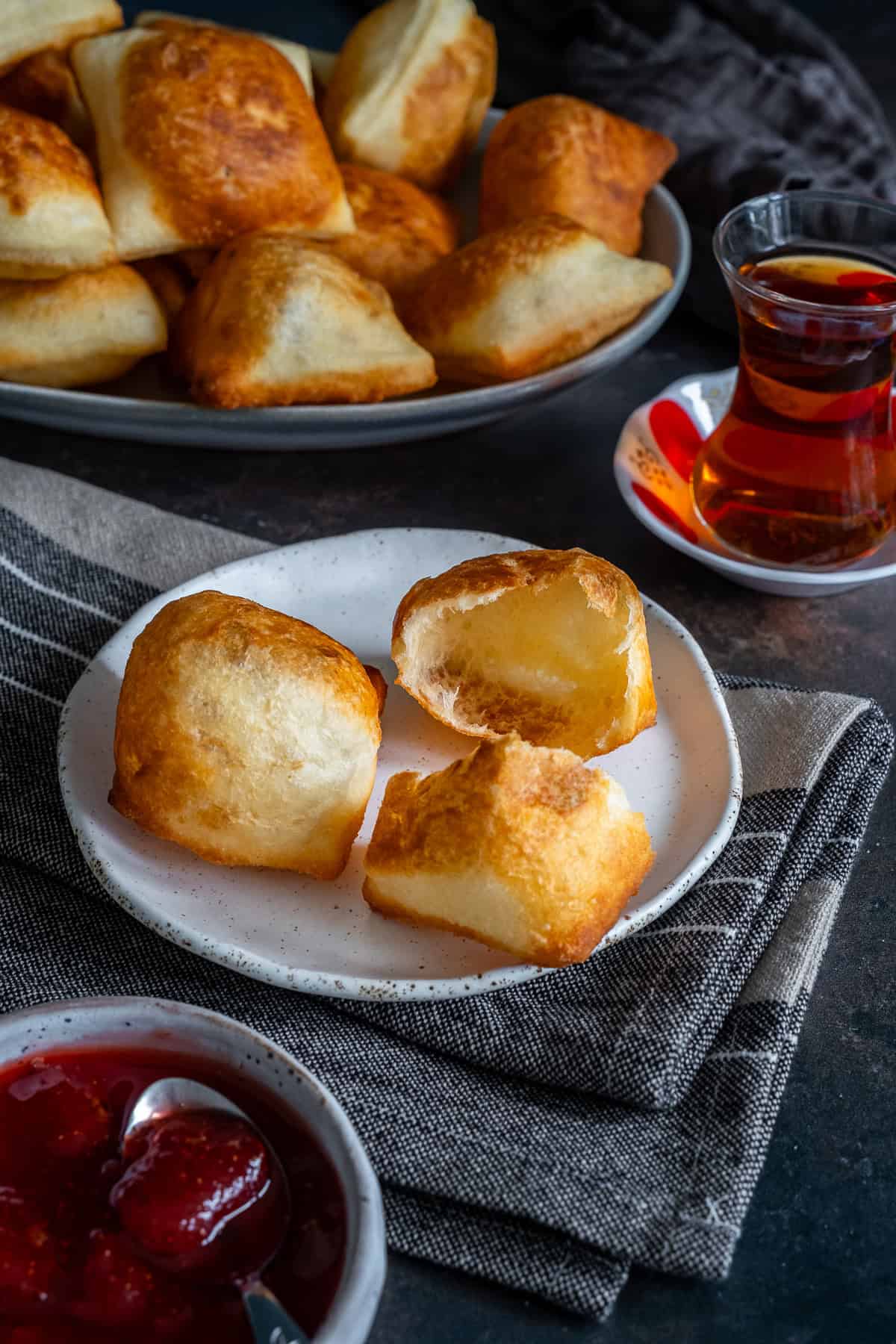 Golden Turkish pişi served with strawberry jam and Turkish tea, with one torn open to show the hollow, airy inside.