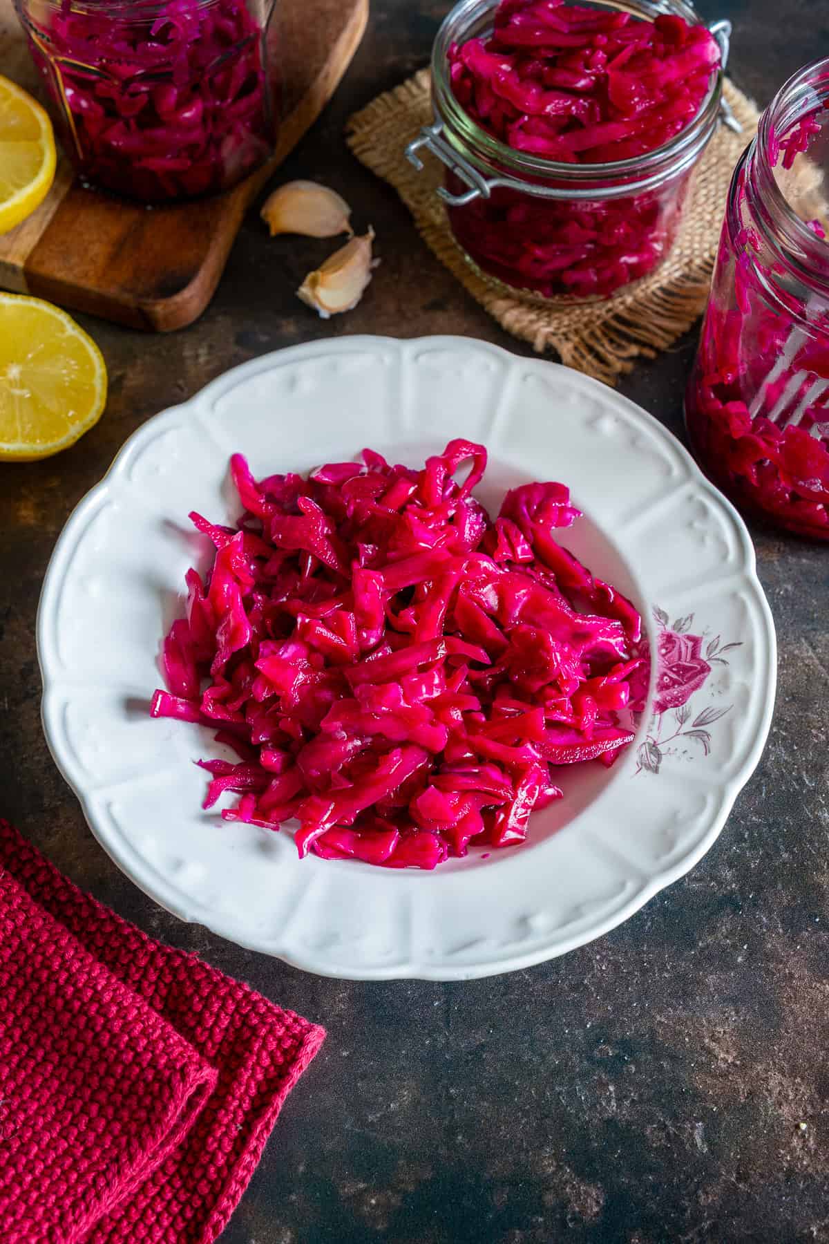 Turkish pickled red cabbage served on a white plate, with jars of pickled cabbage, lemon and garlic in the background.