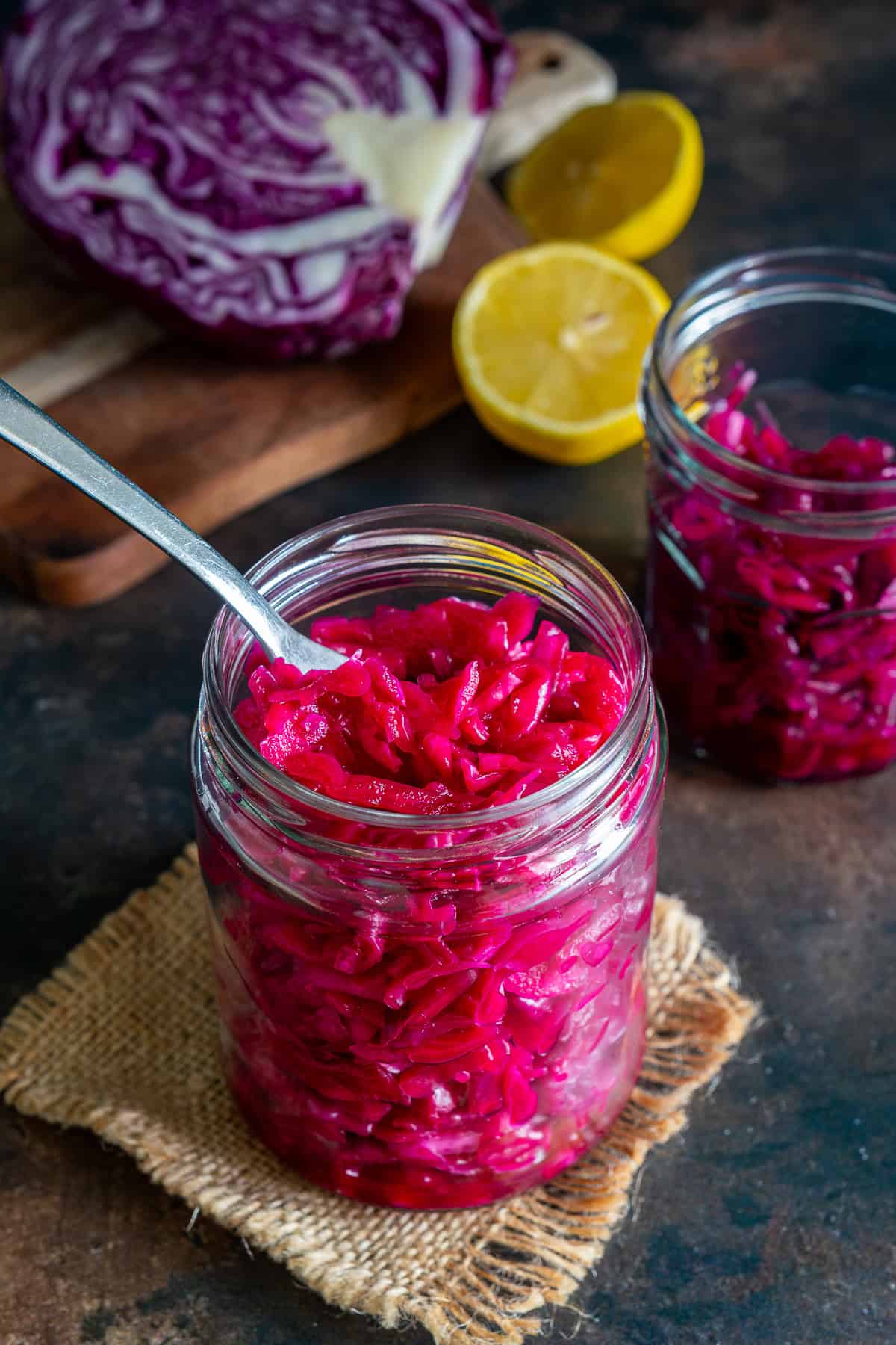 Jar of bright pink Turkish pickled red cabbage with a fork, with red cabbage and lemon in the background.