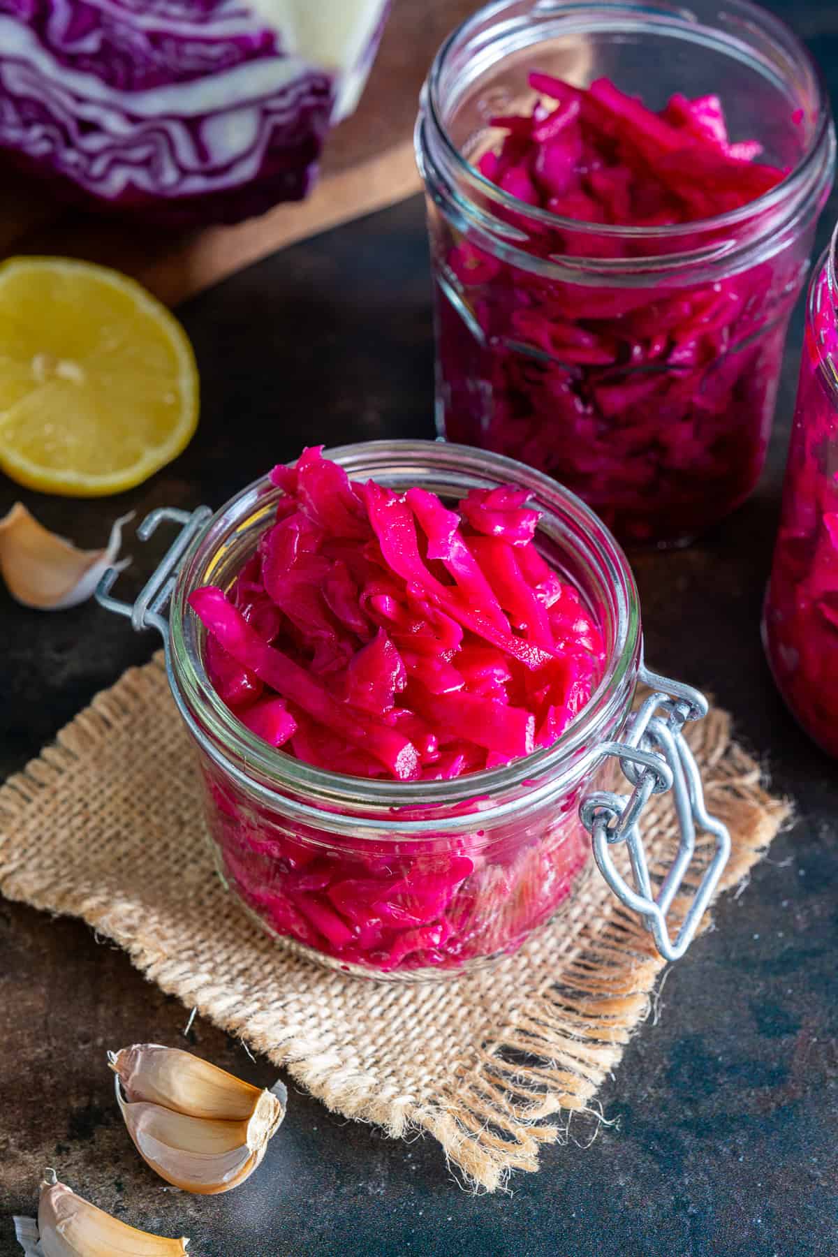 Bright pink Turkish pickled red cabbage in glass jars with lemon, garlic and red cabbage in the background.