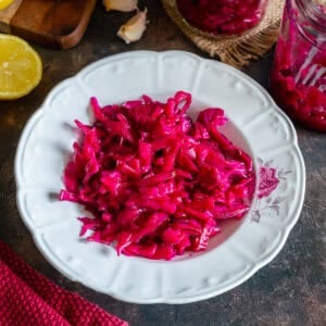 Bright pink Turkish pickled red cabbage served on a white plate, with lemon, garlic and jars of pickled cabbage in the background.