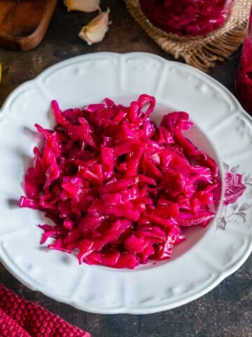 Bright pink Turkish pickled red cabbage served on a white plate, with lemon, garlic and jars of pickled cabbage in the background.