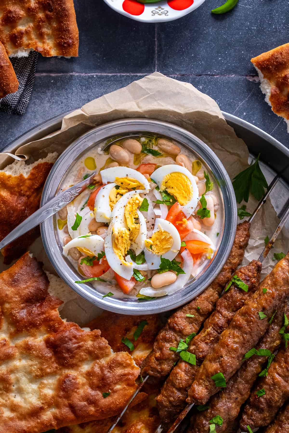 Overhead view of Antalya piyazi (Turkish white bean salad with tahini sauce), topped with hard-boiled eggs, tomatoes, onions and parsley, served with grilled şiş köfte and Turkish bread.
