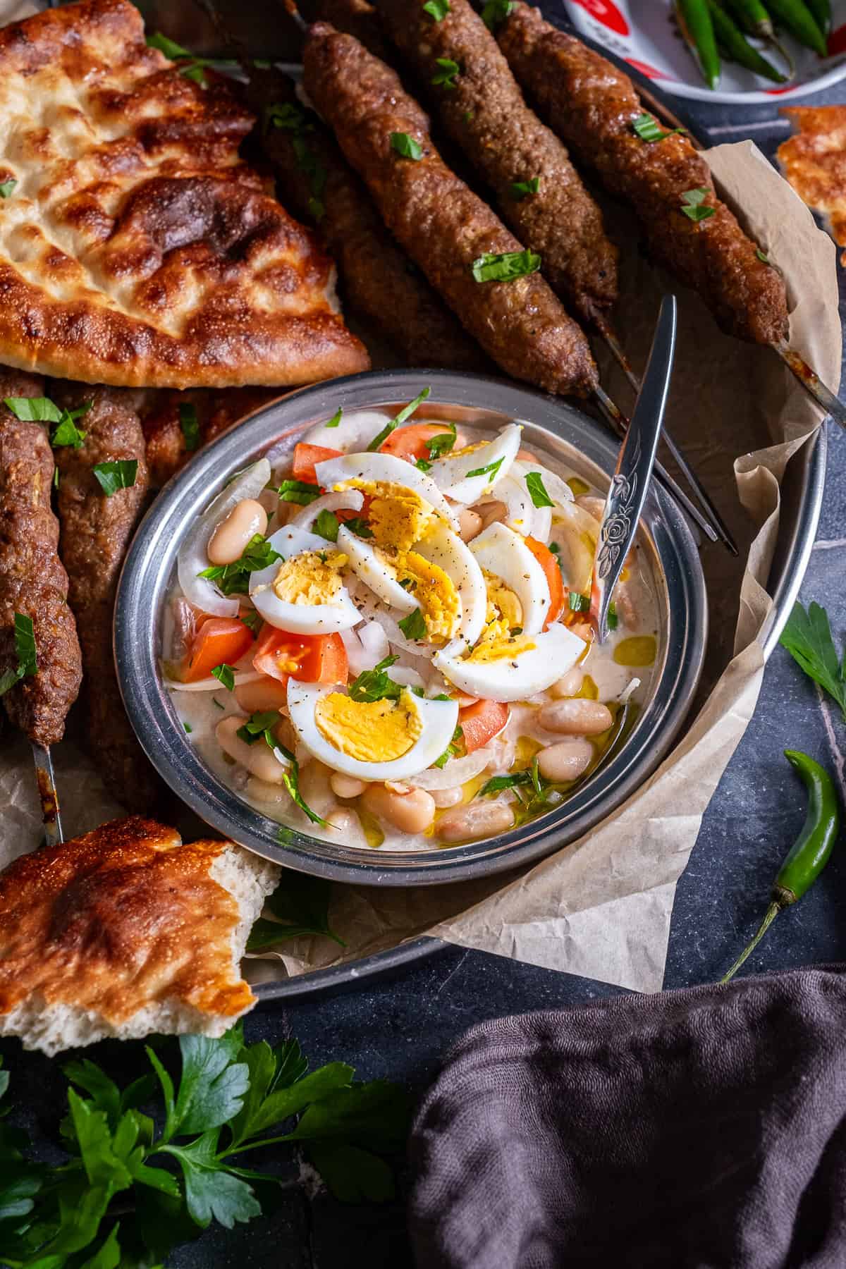 Antalya piyazi topped with sliced hard-boiled eggs, tomatoes, onions and parsley, served alongside grilled şiş köfte, Turkish bread and green peppers.