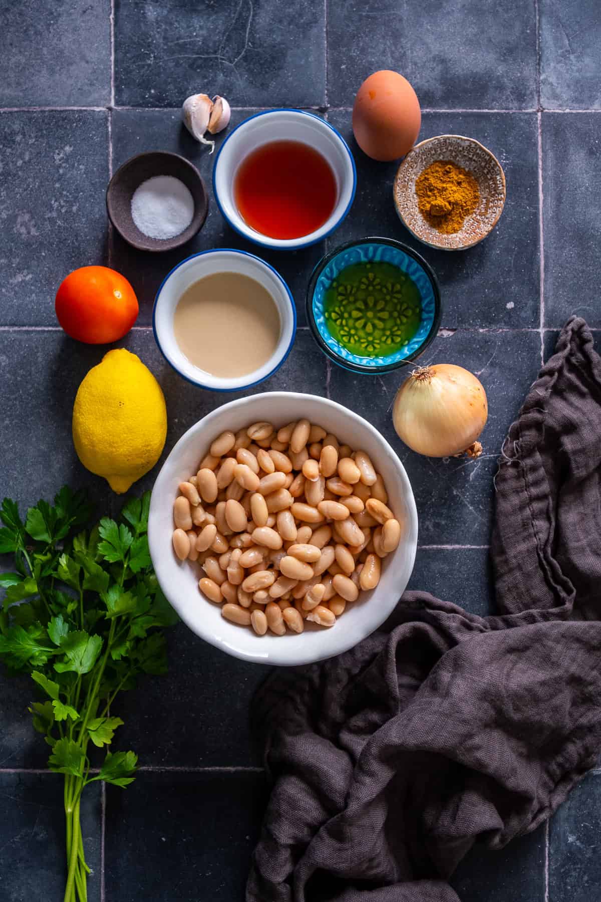 Overhead view of ingredients for Antalya piyazi, including cannellini beans, tahini, vinegar, olive oil, lemon, tomato, onion, parsley, egg, garlic, salt and cumin.