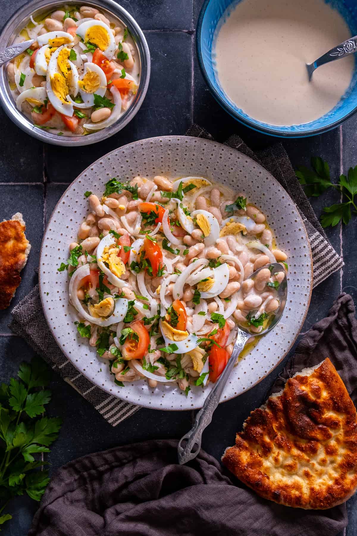 Overhead view of Antalya piyazi served on a large plate with onions, tomatoes, parsley and hard-boiled eggs, alongside a bowl of extra tahini sauce, a smaller portion of piyaz and Turkish bread.