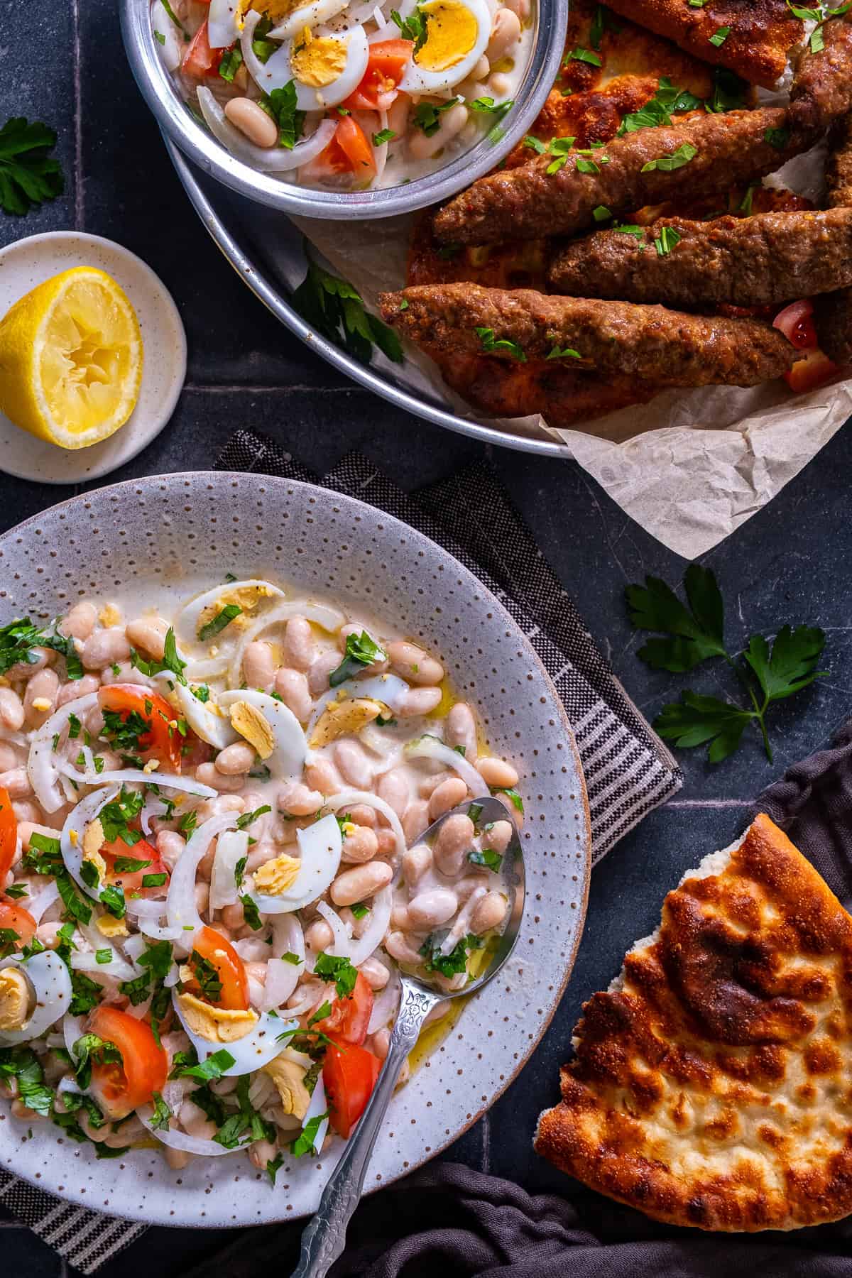 Overhead view of Antalya piyazi topped with hard-boiled eggs, tomatoes, onions and parsley, served alongside grilled şiş köfte and Turkish bread.