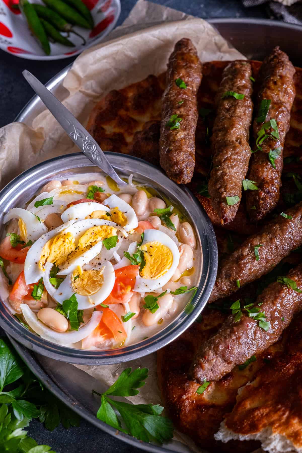 Close-up of Antalya piyazi topped with hard-boiled eggs, tomatoes, onions and parsley, served with grilled şiş köfte and Turkish bread.