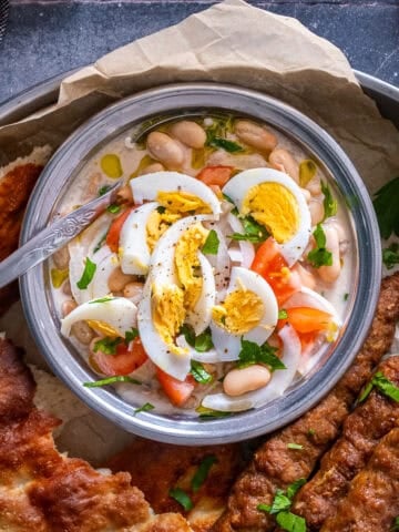 Overhead view of Antalya piyazi topped with hard-boiled eggs, tomatoes, onions and parsley, served with grilled şiş köfte and Turkish bread.