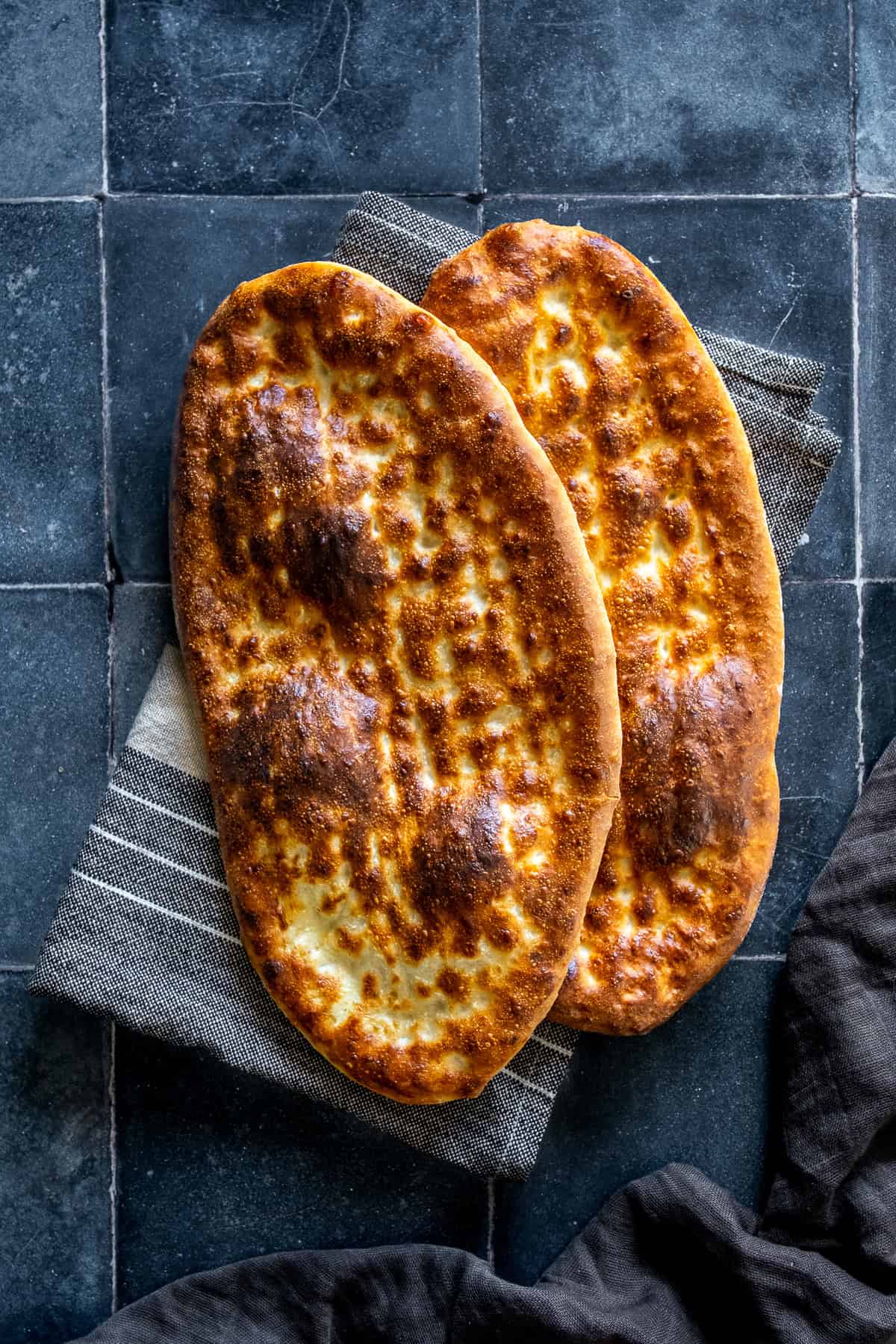 Two freshly baked oval tirnak pides with golden-brown crusts and traditional fingertip patterns, overlapping on a striped kitchen towel.