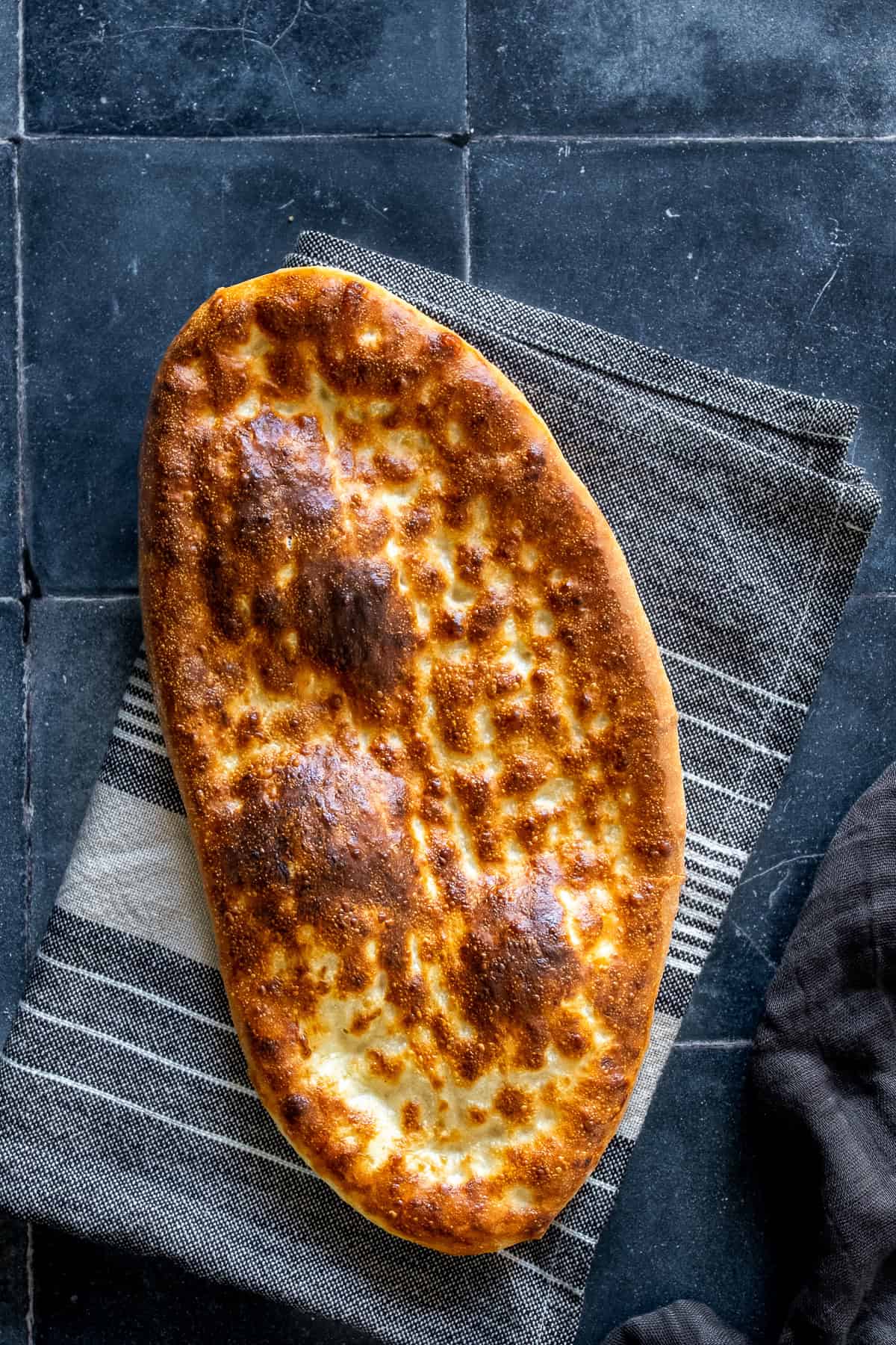 Freshly baked oval tirnak pide with a golden-brown crust and traditional fingertip pattern, resting on a striped kitchen towel.