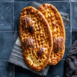 Two freshly baked oval tirnak pides with golden-brown crusts and traditional fingertip patterns, overlapping on a striped kitchen towel.