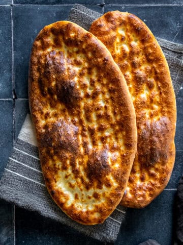 Two freshly baked oval tirnak pides with golden-brown crusts and traditional fingertip patterns, overlapping on a striped kitchen towel.