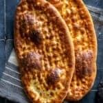 Two freshly baked oval tirnak pides with golden-brown crusts and traditional fingertip patterns, overlapping on a striped kitchen towel.